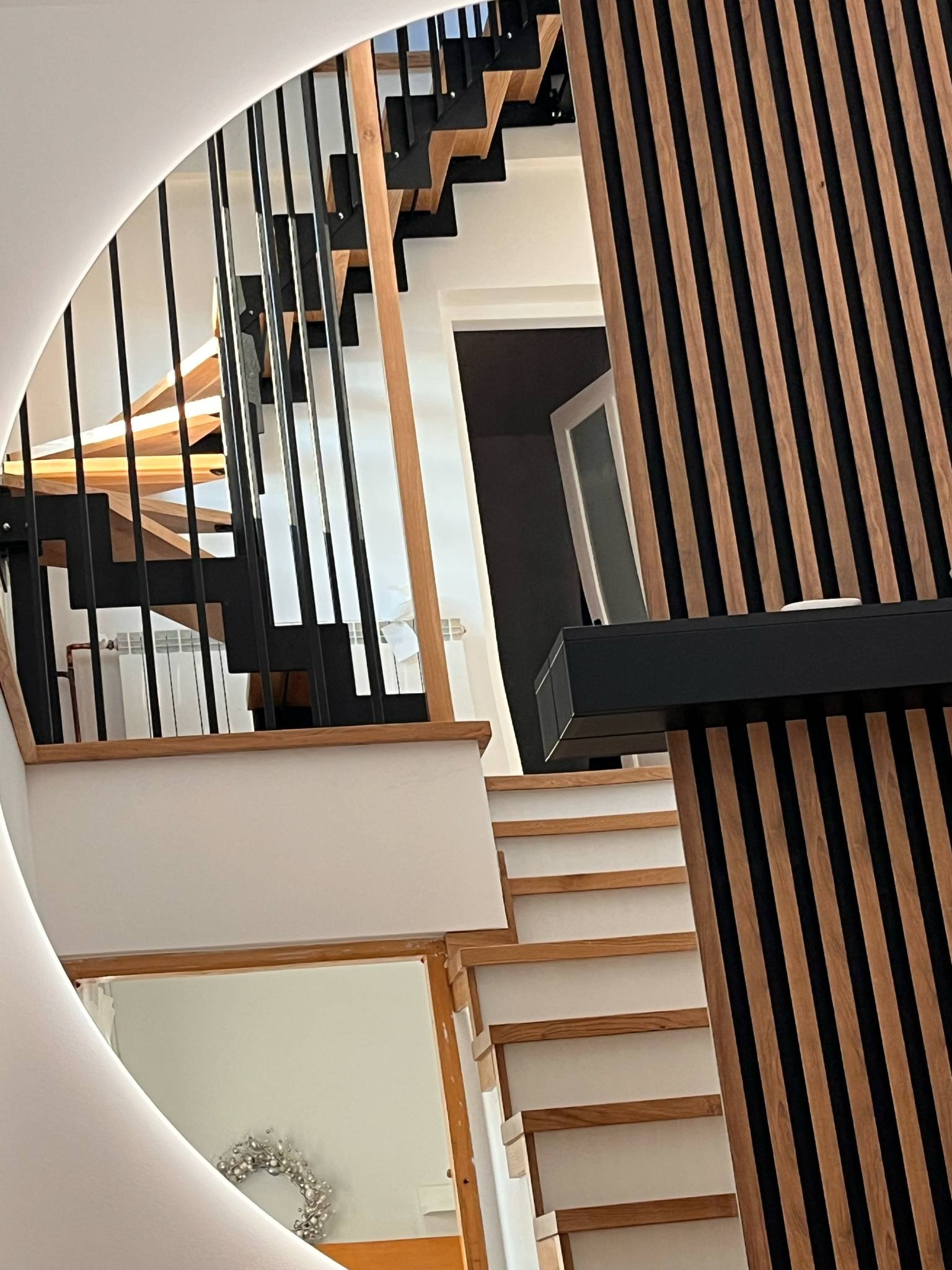 Nowoczesne schody z czarnymi balustradami i drewnianymi stopniami, widoczne przez okrągłe lustro, obok ściana z dekoracyjnych paneli drewnianych.