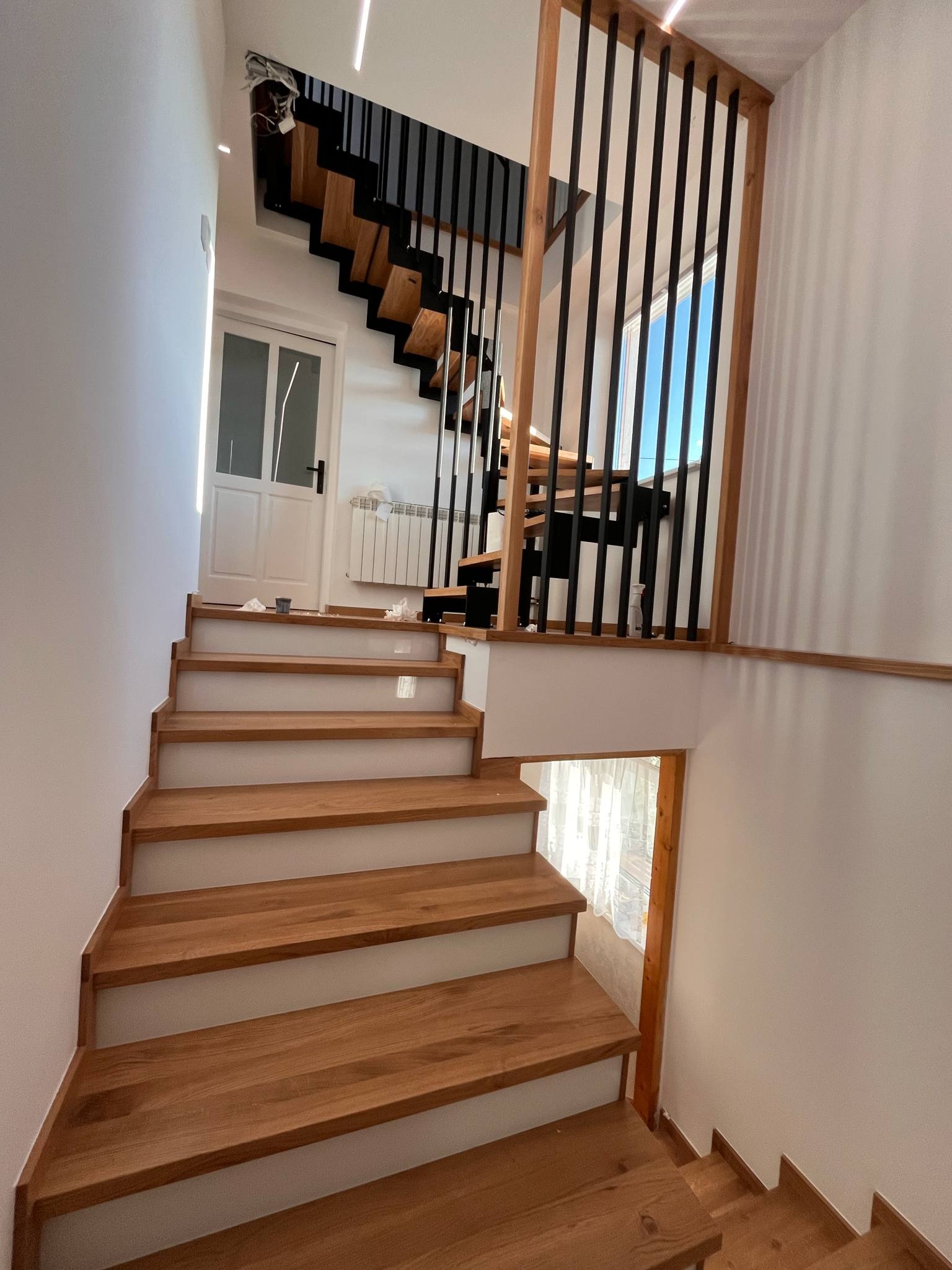 Nowoczesne schody drewniane z czarną balustradą w jasnym wnętrzu. Widoczne drewniane stopnie i fragment dolnych schodów. W tle białe ściany i drzwi.