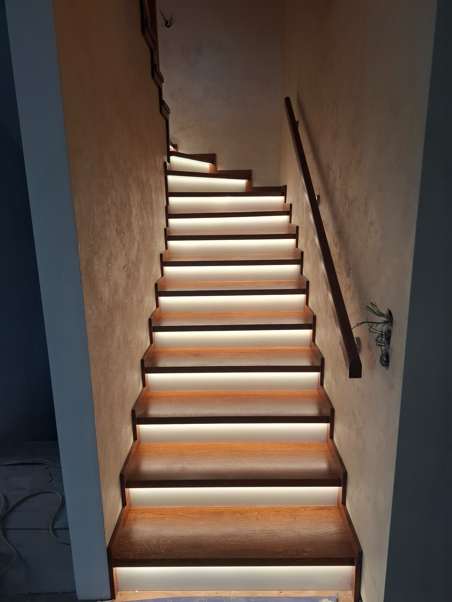 Nowoczesne schody z podświetleniem LED, drewniane stopnie i minimalistyczna balustrada. Widoczne niedokończone prace elektryczne przy ścianie.