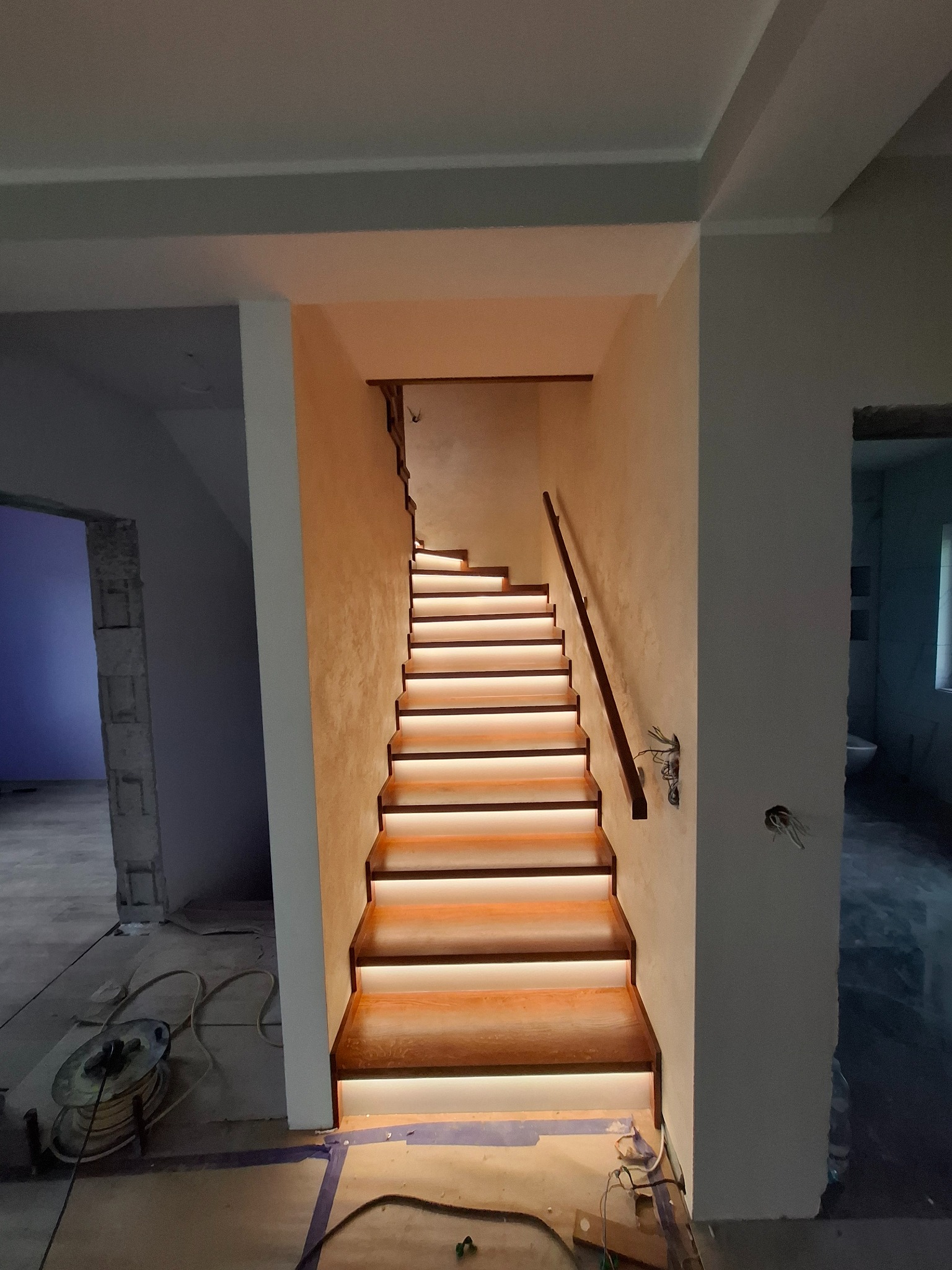 Nowoczesne schody z podświetleniem LED, drewniane stopnie i poręcz, widok z dołu na górę, surowy stan wykończenia wnętrza.