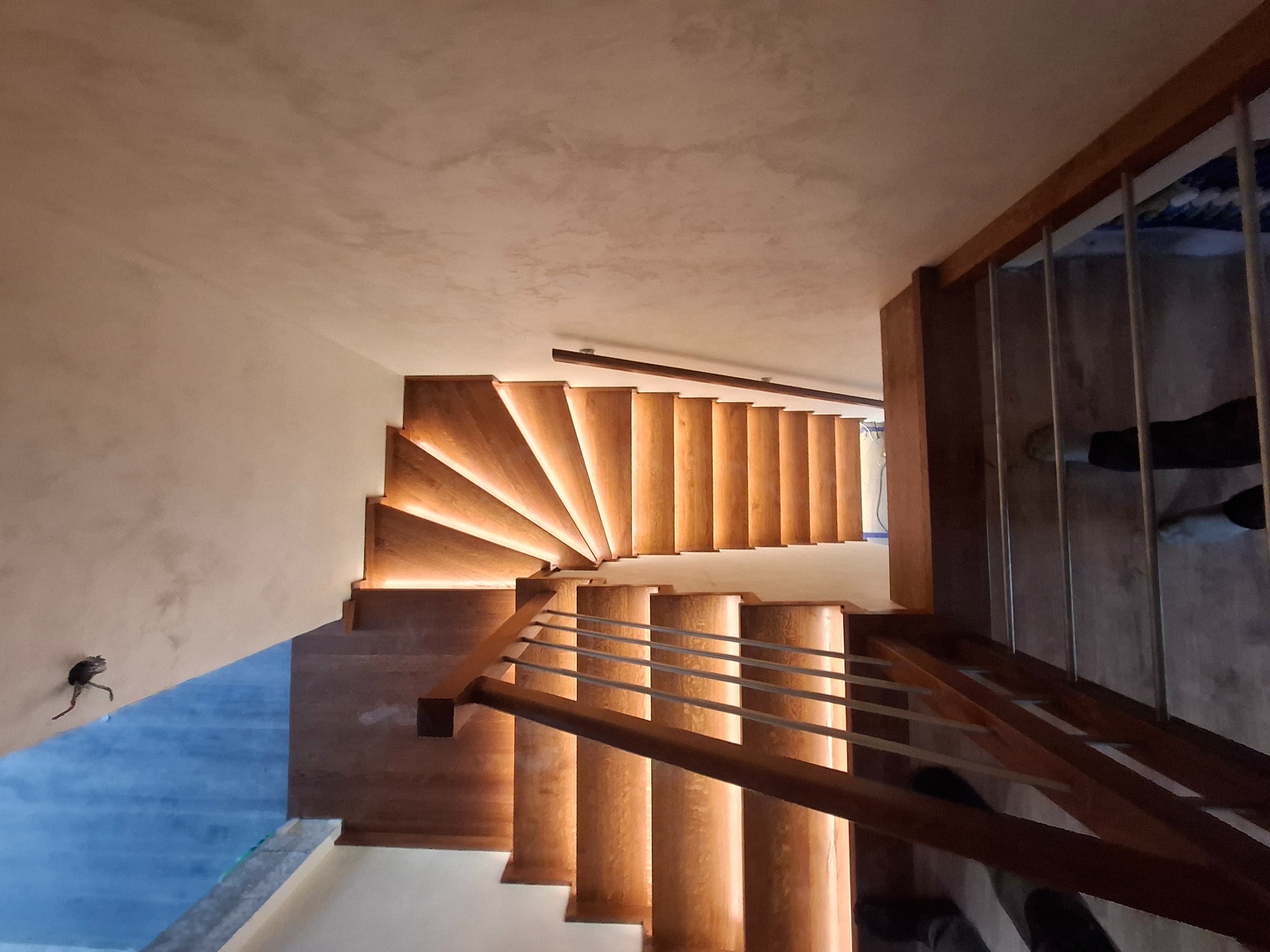 Kaskadowe schody z podświetleniem LED na stopniach, widziane z góry, z drewnianą balustradą o metalowych prętach. Neutralne tło ścian i sufitu.