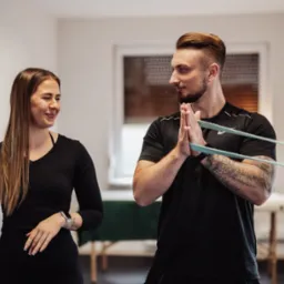 Mężczyzna z tatuażami na ramieniu ćwiczy z gumą oporową, obok uśmiechnięta kobieta w czarnym stroju sportowym, jasne wnętrze z oknem.