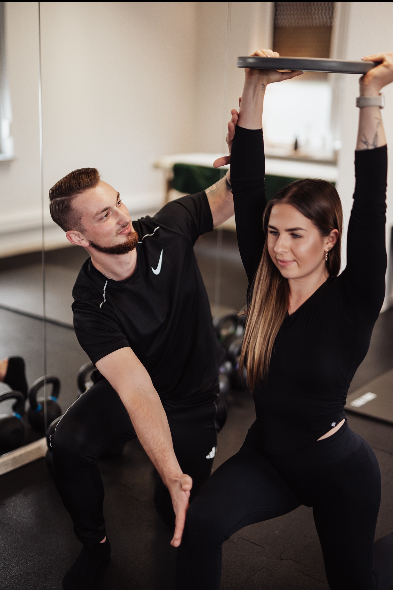Kobieta w czarnym stroju sportowym ćwiczy przysiady z obciążeniem trzymanym nad głową, asystuje jej mężczyzna w sportowym ubraniu w siłowni z lustrami i kettlebells w tle.