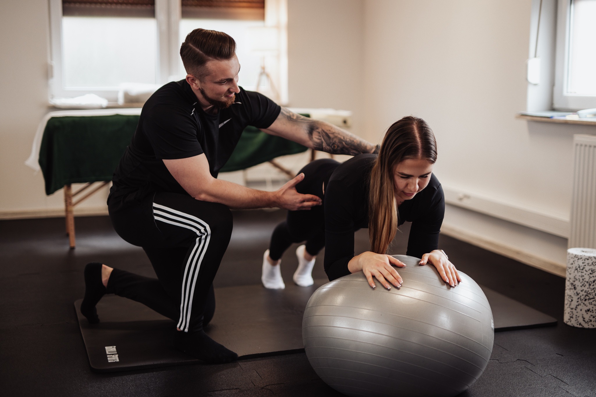 Kobieta w pozycji plank na piłce gimnastycznej, nadzorowana przez trenera z Wrocławia, który koryguje jej postawę. Widoczne tatuaże na ramieniu trenera i mata do ćwiczeń.