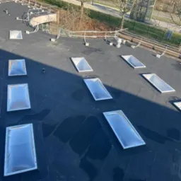 Czarny dach płaski z zamontowanymi świetlikami dachowymi o prostokątnym kształcie, widok z góry, w tle drzewa i budynki.