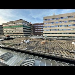 Dekarstwo Paluszczak - Rozległy dach budynku komercyjnego z widocznymi ekipami i materiałami przygotowanymi do prac dekarskich, z perspektywy dachu innego budynku.