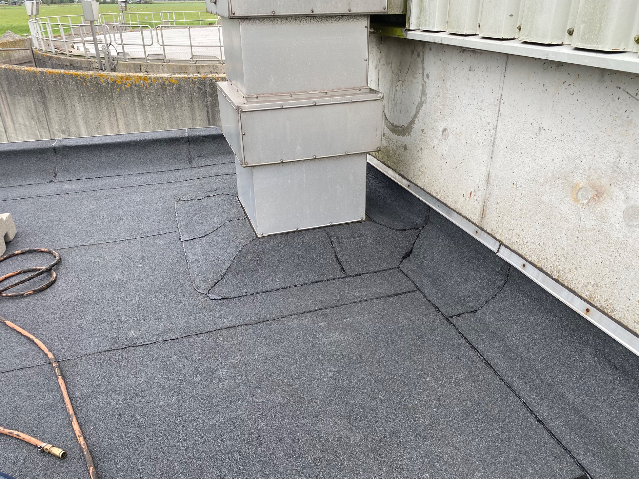 Płaski dach pokryty papą z widocznymi łączeniami, obok metalowa konstrukcja wentylacyjna i fragment betonowej ściany, w rogu leży zwinięty wąż.
