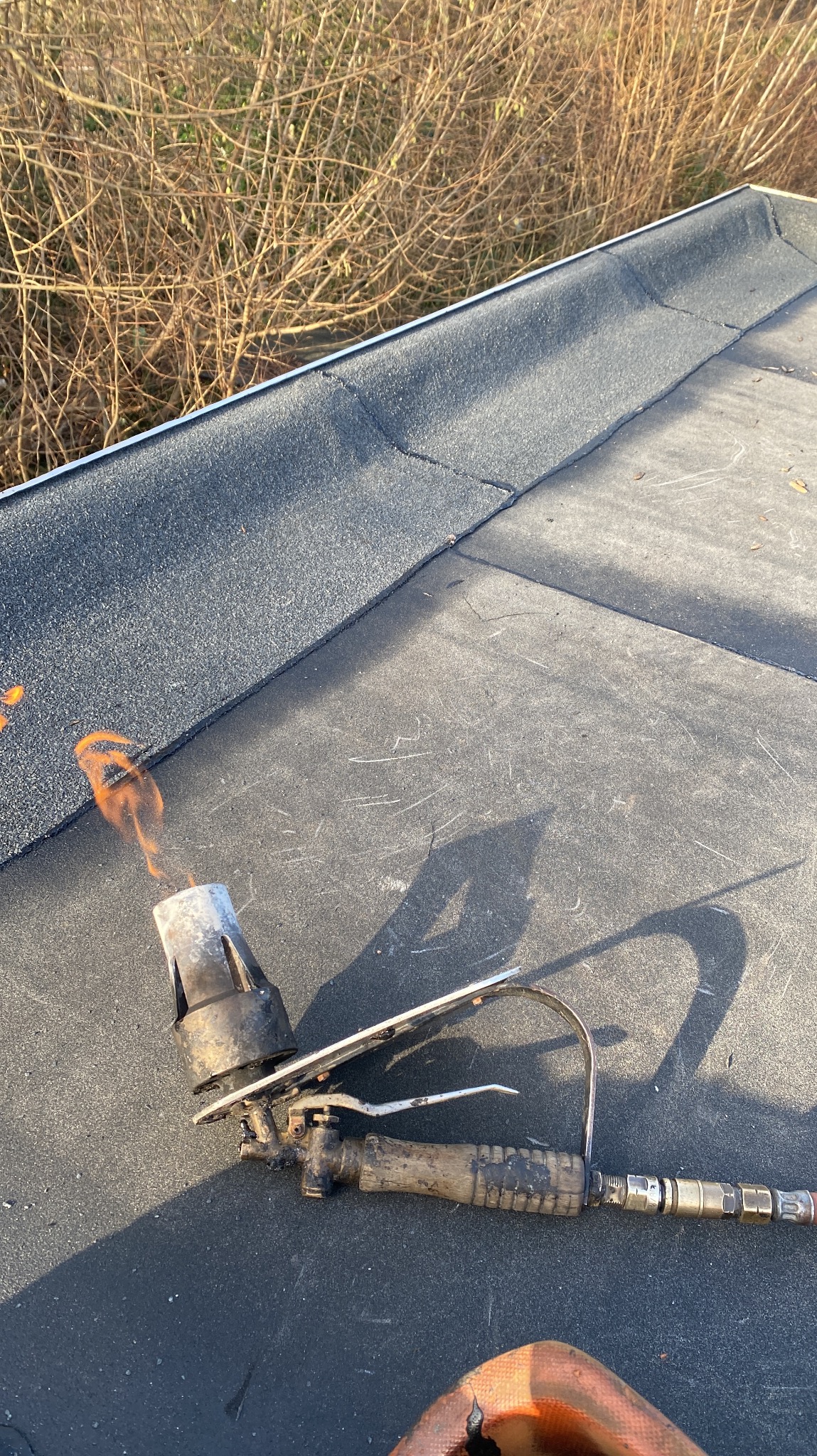 Palnik dekarski w trakcie pracy na płaskim dachu pokrytym papą, widoczne płomienie i cień narzędzia na tle krzewów.