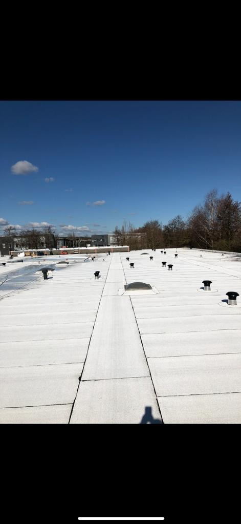 Widok z góry na płaski, biały dach pokryty papą termozgrzewalną z licznymi wywietrznikami i świetlikiem, na tle błękitnego nieba i zabudowań miejskich. Widoczny cień osoby robiącej zdjęcie.
