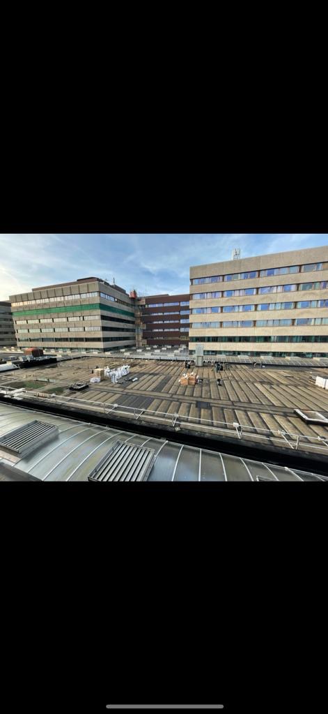 Rozległy dach budynku komercyjnego z widocznymi ekipami i materiałami przygotowanymi do prac dekarskich, z perspektywy dachu innego budynku.