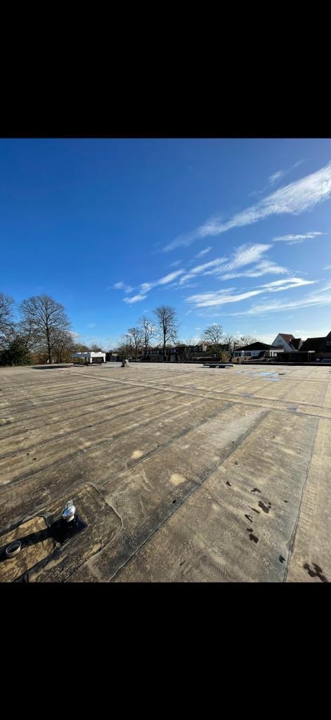 Płaski, gumowy dach z widocznymi łączeniami i elementami mocującymi, sfotografowany pod błękitnym niebem z lekkimi chmurami, w tle widoczne drzewa i budynki.