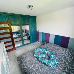 Roman Idczak - Sypialnia po odświeżeniu: turkusowa szafa wnękowa z lustrem, łóżko z szarą pościelą w liście i poduszką w tropikalne wzory, panelowy zagłówek w odcieniach niebieskiego i fioletu.