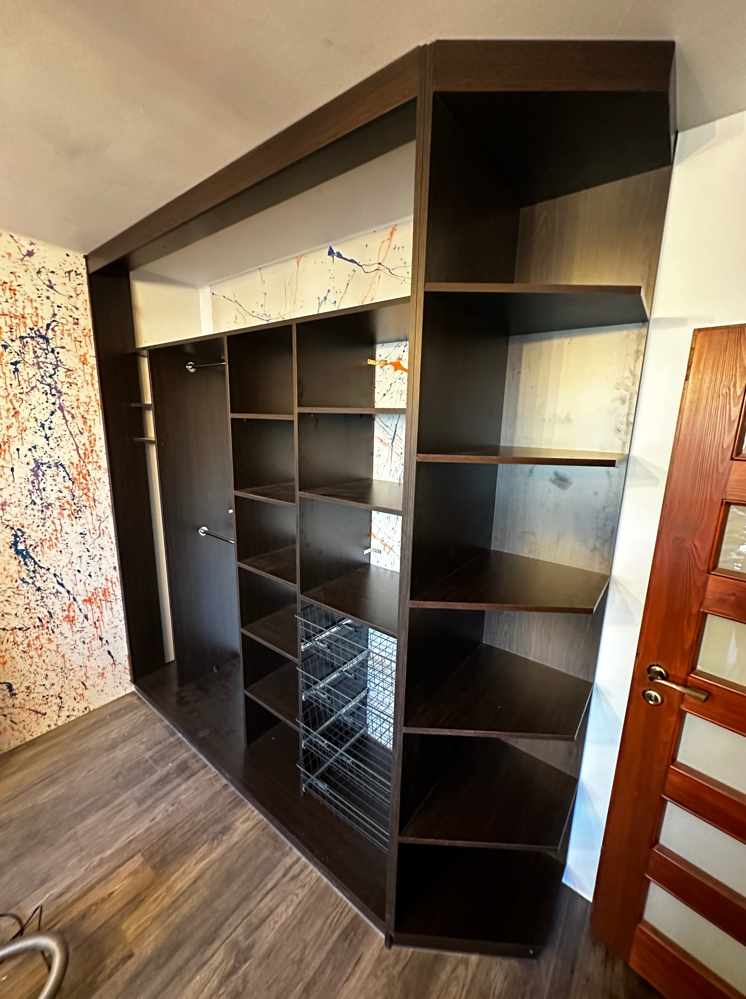 Ciemnobrązowa, narożna garderoba z metalowymi koszami, zamontowana w pokoju z podłogą imitującą drewno i ścianą z abstrakcyjnymi plamami farby.