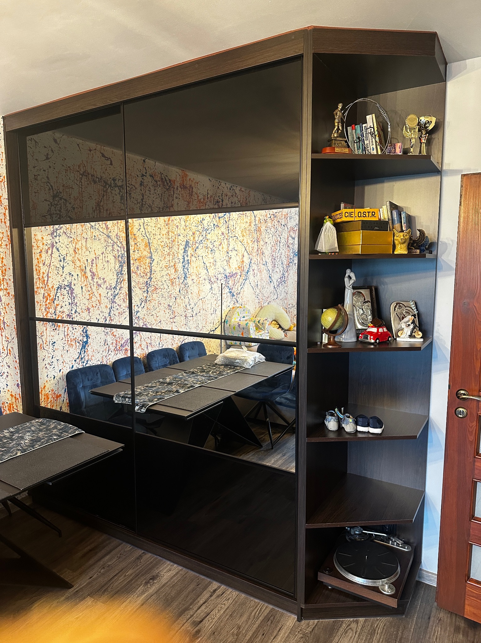 Szafa wnękowa z przesuwnymi drzwiami lustrzanymi, zintegrowana z regałem narożnym, na którym znajdują się dekoracje i sprzęt audio, odbijająca stół i krzesła na tle ściany z abstrakcyjnym malowaniem.