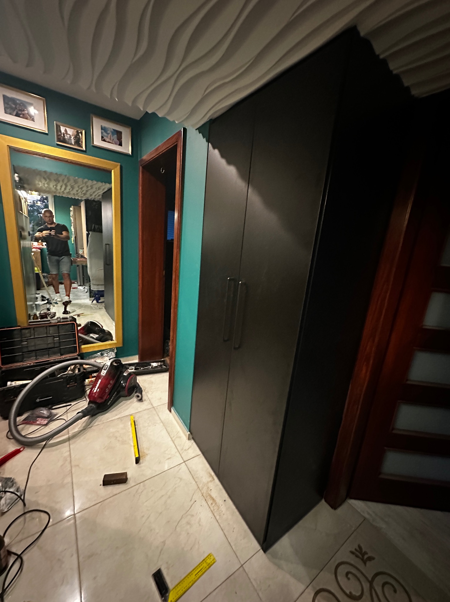 Montaż czarnej szafy wnękowej na tle turkusowej ściany, widoczny odkurzacz, narzędzia i odbicie osoby w lustrze z żółtą ramą.