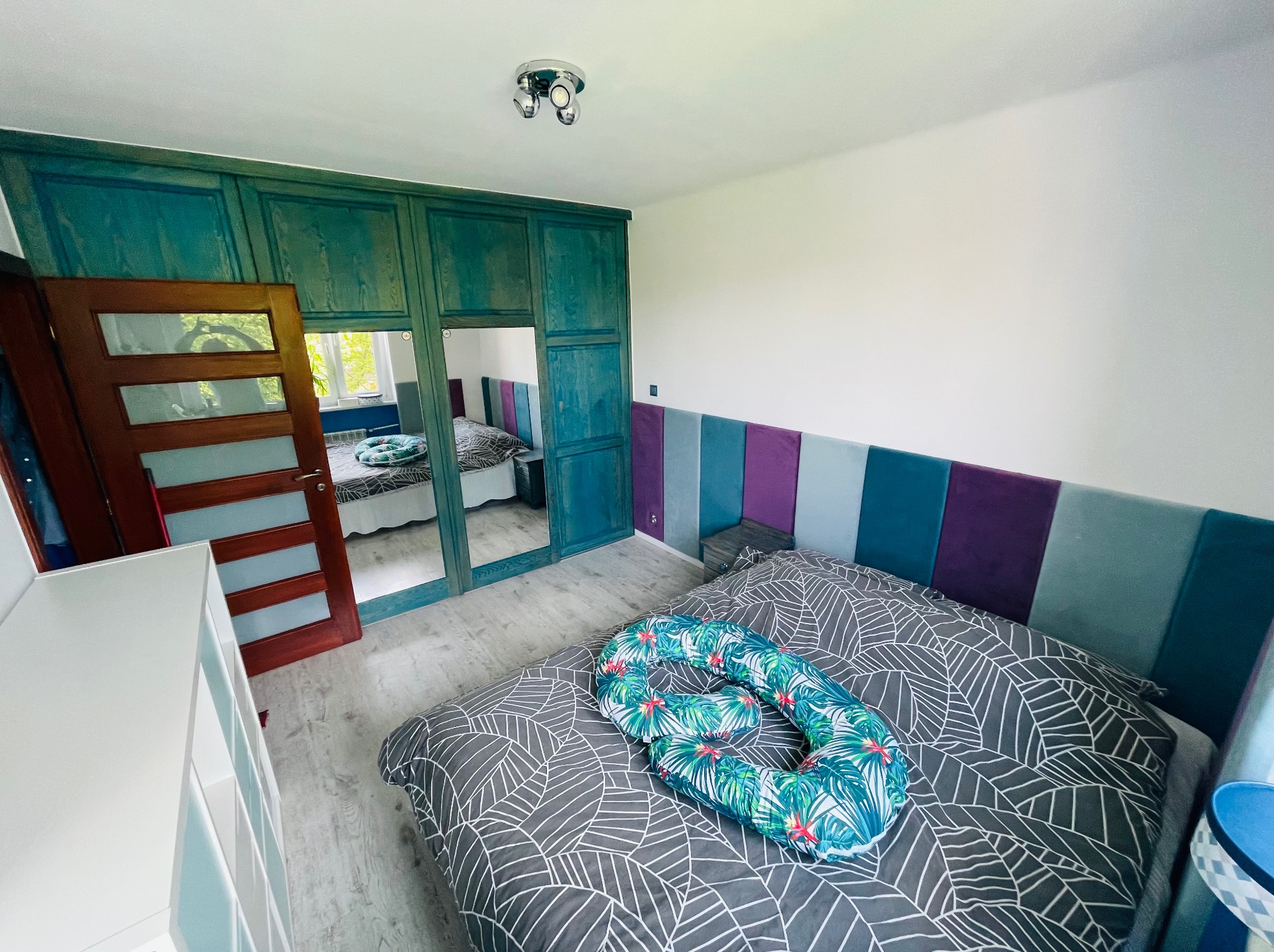 Sypialnia po odświeżeniu: turkusowa szafa wnękowa z lustrem, łóżko z szarą pościelą w liście i poduszką w tropikalne wzory, panelowy zagłówek w odcieniach niebieskiego i fioletu.