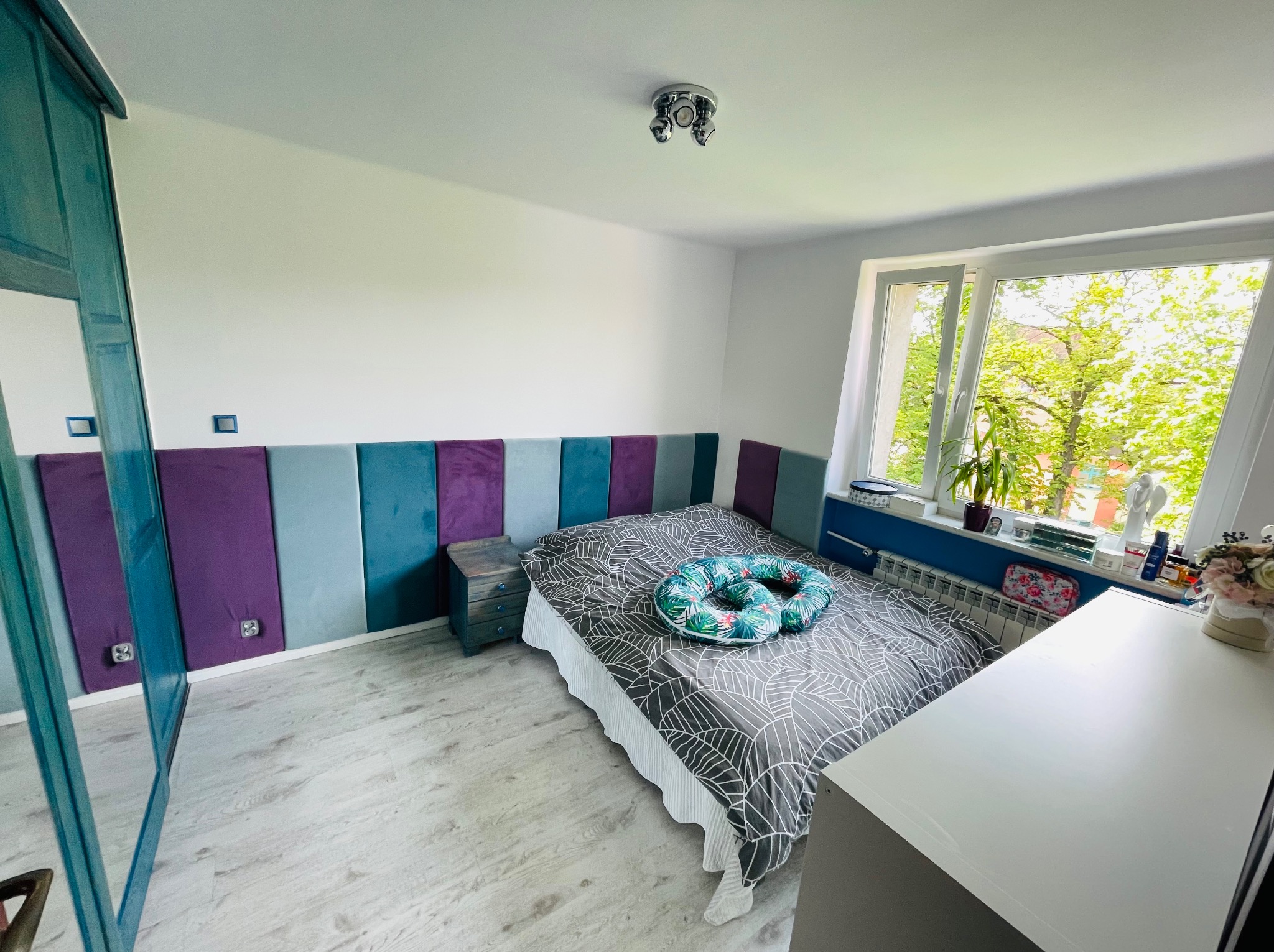 Sypialnia z panelami ściennymi w odcieniach fioletu i turkusu, łóżko z szarą pościelą w geometryczne wzory oraz poduszką w tropikalne liście, drewniana podłoga i widok z okna na zieleń drzew.