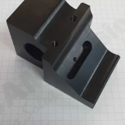 Precyzyjnie wykonany, czarny element z metalu z widocznymi otworami i gwintami, leżący na papierze w kratkę, z logotypem firmy Mdivision w tle.