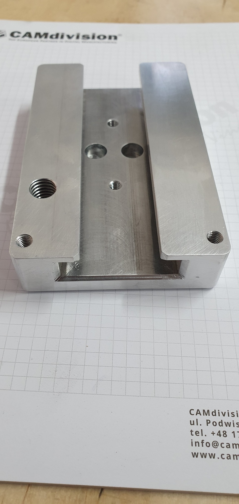 Metalowy element z gwintowanymi otworami, prawdopodobnie uchwyt lub podstawa, wykonany w procesie obróbki w Wrzosowej, leżący na papierze w kratkę.