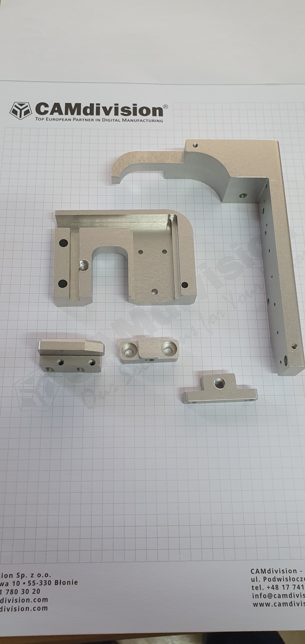 Precyzyjnie obrobione, srebrne elementy metalowe z widocznymi otworami, ułożone na papierze w kratkę z logo CAMdivision, ukazujące detale wykonania.