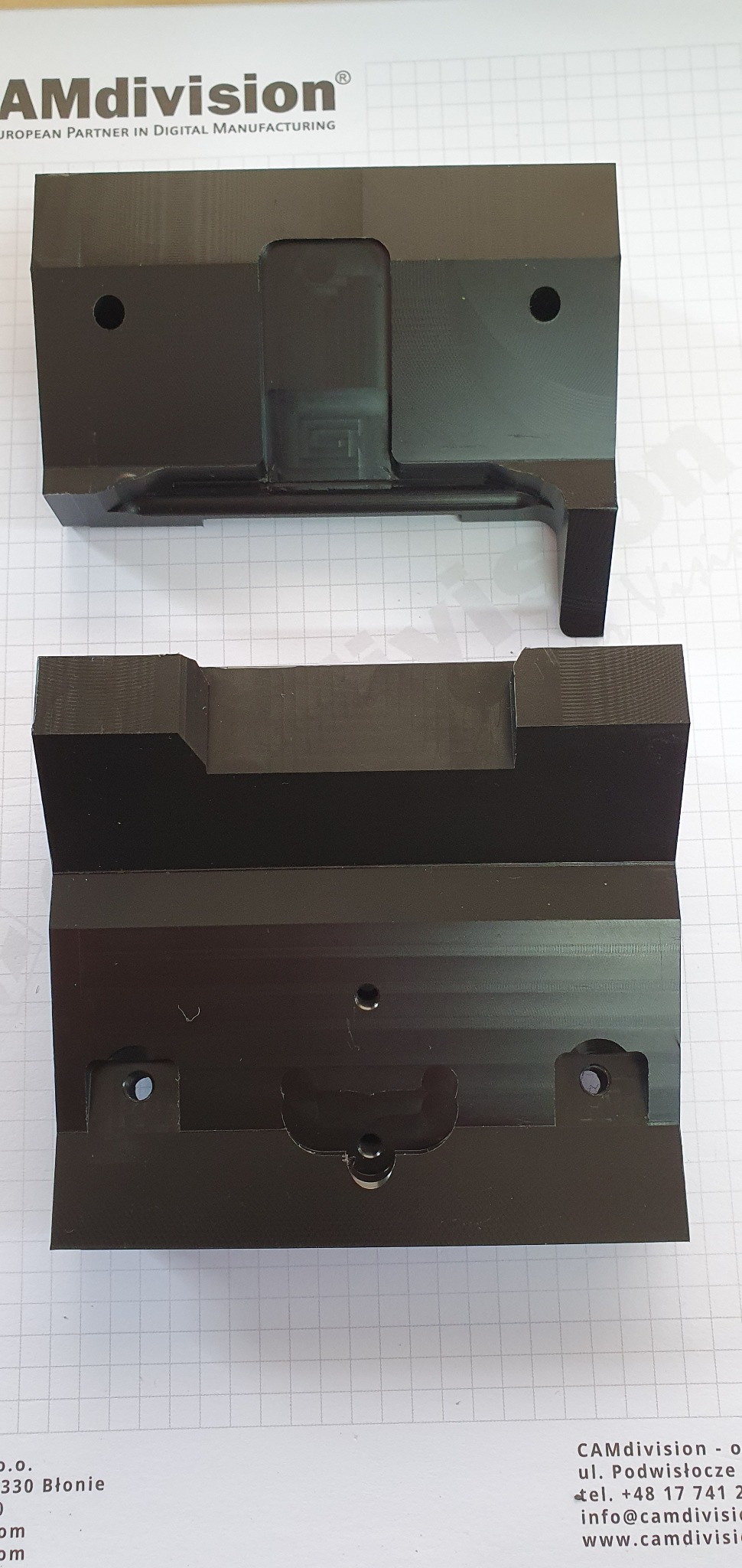 Dwie czarne, precyzyjnie wyfrezowane, identyczne części z metalu leżące na papierze w kratkę, widoczne otwory i wgłębienia.