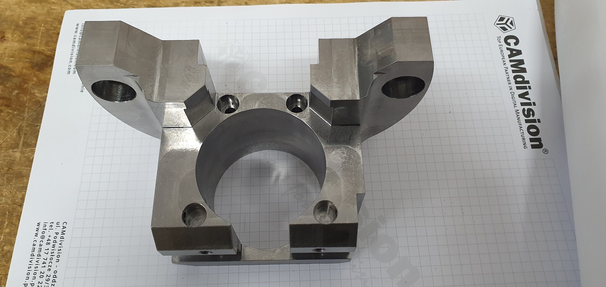 Metalowy element konstrukcyjny z otworami, obrobiony na gładko, leżący na kartce papieru z logotypem firmy CAMdivision.
