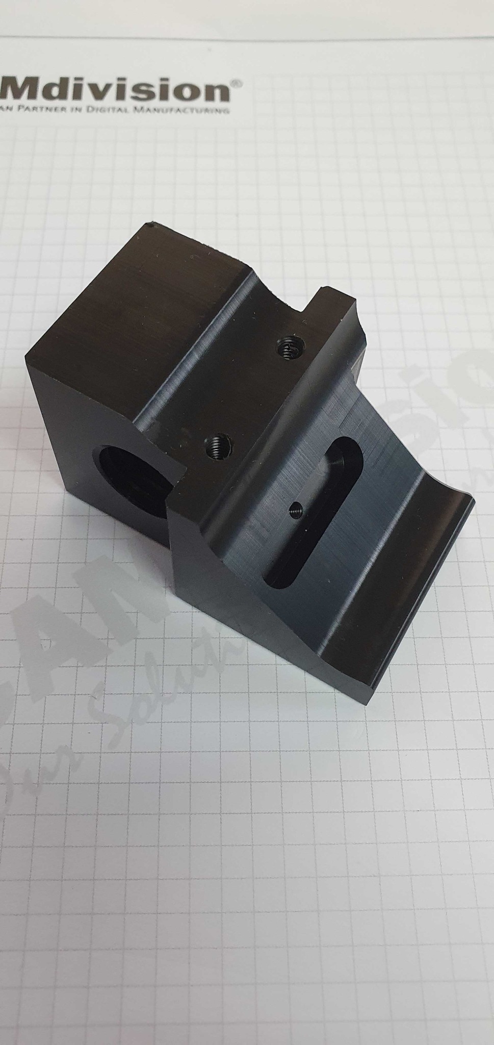 Precyzyjnie wykonany, czarny element z metalu z widocznymi otworami i gwintami, leżący na papierze w kratkę, z logotypem firmy Mdivision w tle.