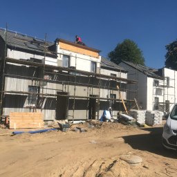 BP Spolka z OO - Budynek w trakcie prac elewacyjnych, z widocznymi rusztowaniami, ociepleniem i pracownikiem w czerwonej koszulce na dachu, na pierwszym planie plac budowy z piachem i materiałami budowlanymi.