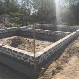 Fundamenty domu w trakcie budowy, widoczne bloczki betonowe i piasek wewnątrz, teren niezagospodarowany, w tle drzewa.