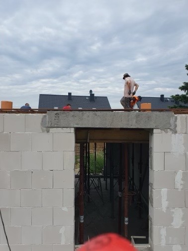 Wznoszenie ściany z betonu komórkowego na budowie domu jednorodzinnego, widoczne szalunki i zbrojenie nad otworem okiennym, pracownicy w tle.