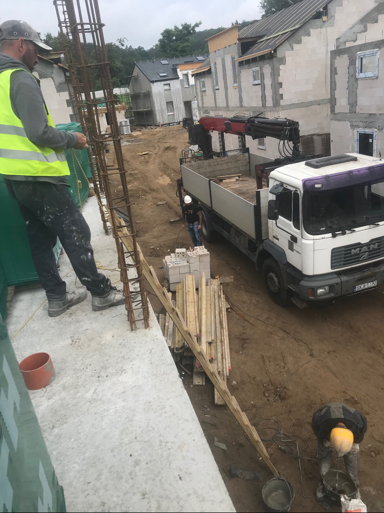 Budowa domu w trakcie realizacji: pracownik wiąże zbrojenie na fundamencie, w tle widać kolejne budynki w stanie surowym, ciężarówka z dźwigiem i pracownik przygotowujący zaprawę.