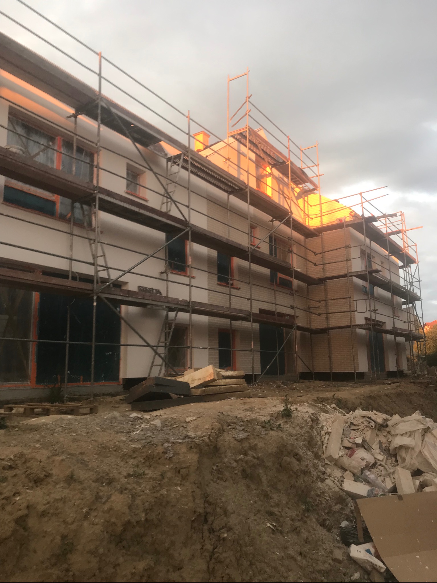 Budynek wielorodzinny w trakcie ocieplania elewacji, widoczne rusztowania, częściowo otynkowana fasada i fragmenty ceglanej ściany, teren budowy z gruzem.