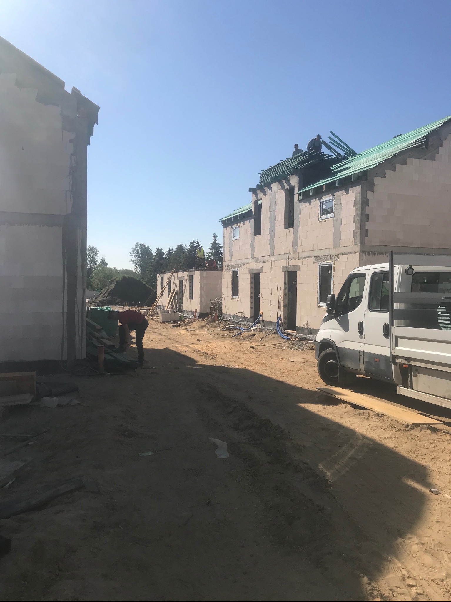 Budowa domu w Gdańsku: robotnicy pracują na dachu, układając konstrukcję, a inny pracownik przenosi materiały budowlane obok zaparkowanego samochodu dostawczego.