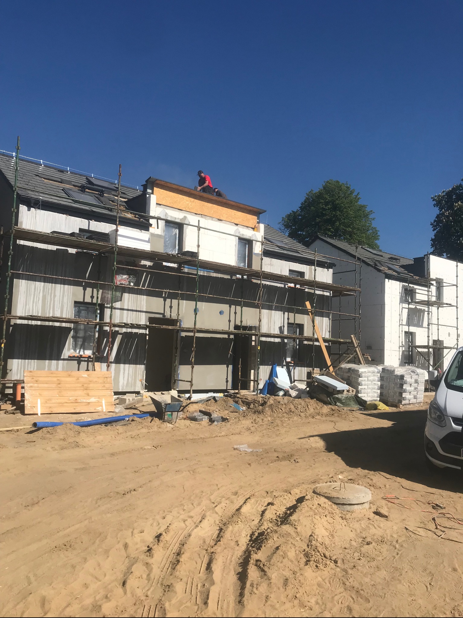 Budynek w trakcie prac elewacyjnych, z widocznymi rusztowaniami, ociepleniem i pracownikiem w czerwonej koszulce na dachu, na pierwszym planie plac budowy z piachem i materiałami budowlanymi.
