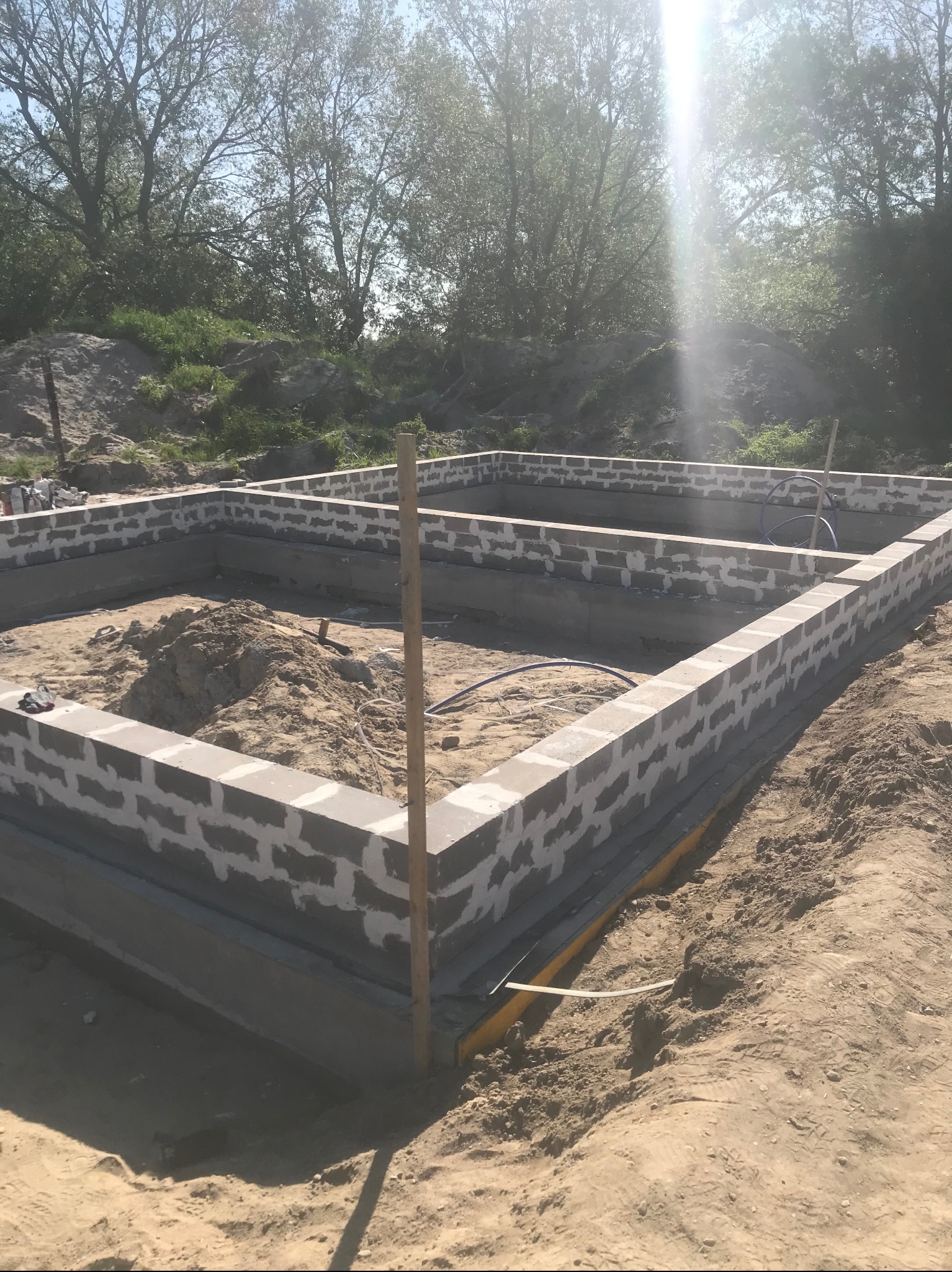 Fundamenty domu w trakcie budowy, widoczne bloczki betonowe i piasek wewnątrz, teren niezagospodarowany, w tle drzewa.
