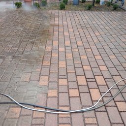 Marcel Mazurkiewicz - Częściowo wyczyszczona kostka brukowa, widoczna różnica w kolorze i stopniu zabrudzenia, z przewodami węży na pierwszym planie i roślinnością w tle.