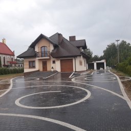 Marcel Mazurkiewicz - Nowo ułożona, mokra, grafitowa kostka brukowa z białymi, krętymi wzorami na podjeździe przed domem z brązowym dachem, widok na kościół w tle.