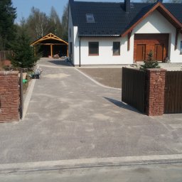Marcel Mazurkiewicz - Nowo ułożona, szara kostka brukowa prowadząca do nowoczesnego domu z czarnym dachem i drewnianą bramą wjazdową, widoczne narzędzia i materiały budowlane.
