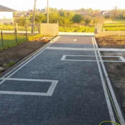 Nowo ułożona, szara kostka brukowa z białymi wstawkami w kształcie prostokątów, obrzeża z kostki, widoczne przygotowane miejsca na zieleń, w tle dom i ogród.