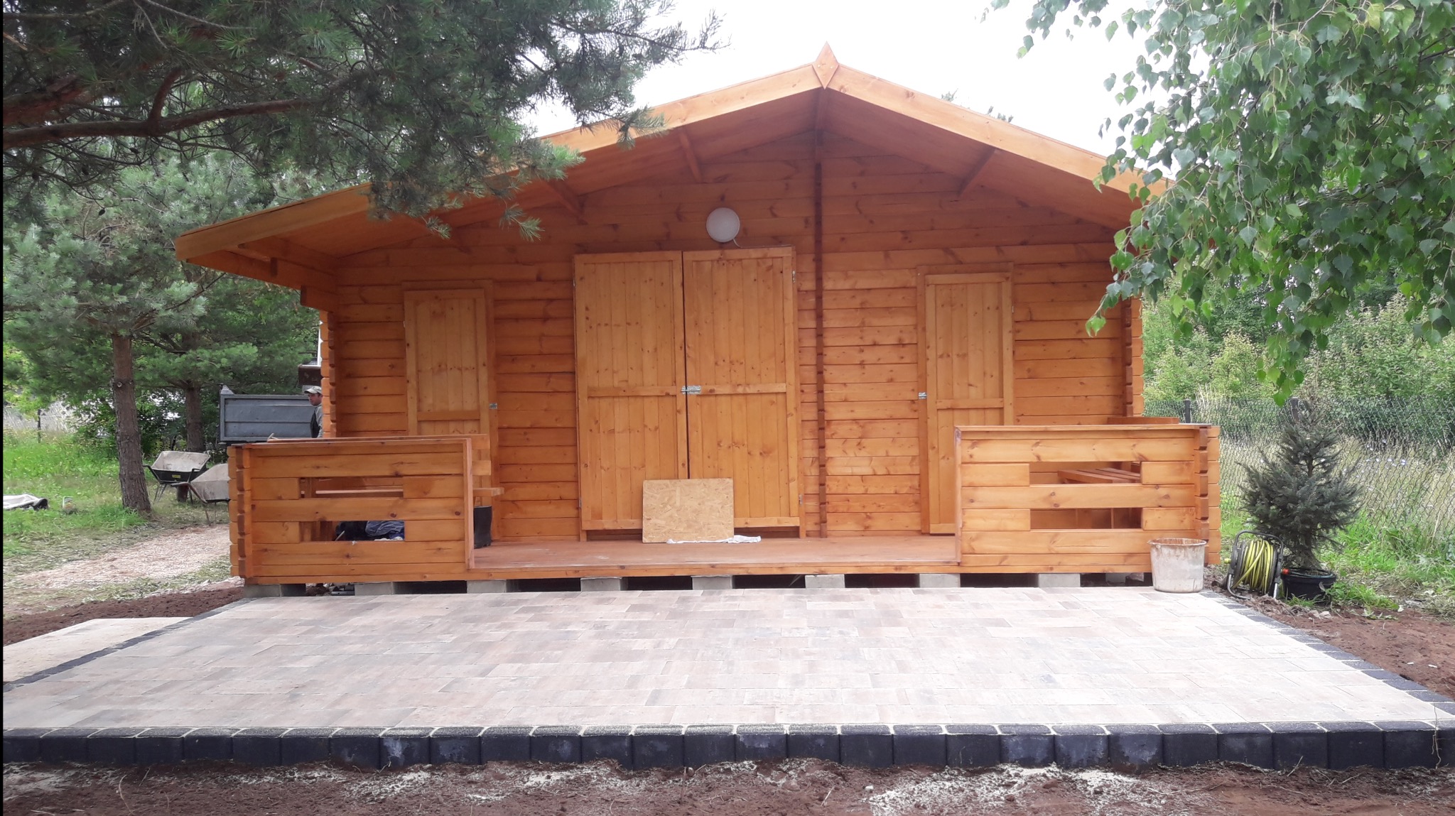 Nowo postawiony drewniany domek narzędziowy z dwuskrzydłowymi drzwiami na betonowej podmurówce, z wybrukowanym tarasem z kostki w odcieniach beżu z czarną obwódką, na tle zieleni.