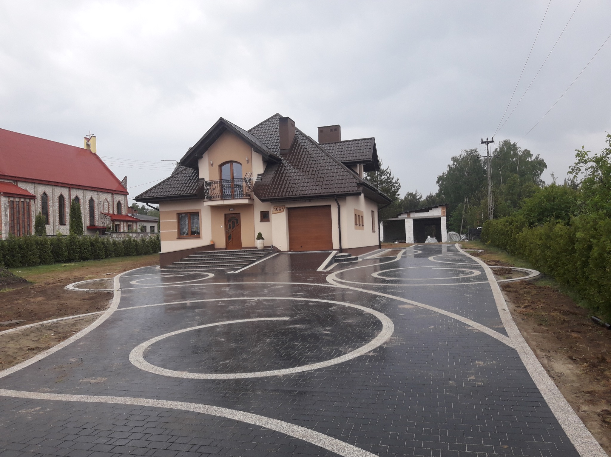 Nowo ułożona, mokra, grafitowa kostka brukowa z białymi, krętymi wzorami na podjeździe przed domem z brązowym dachem, widok na kościół w tle.