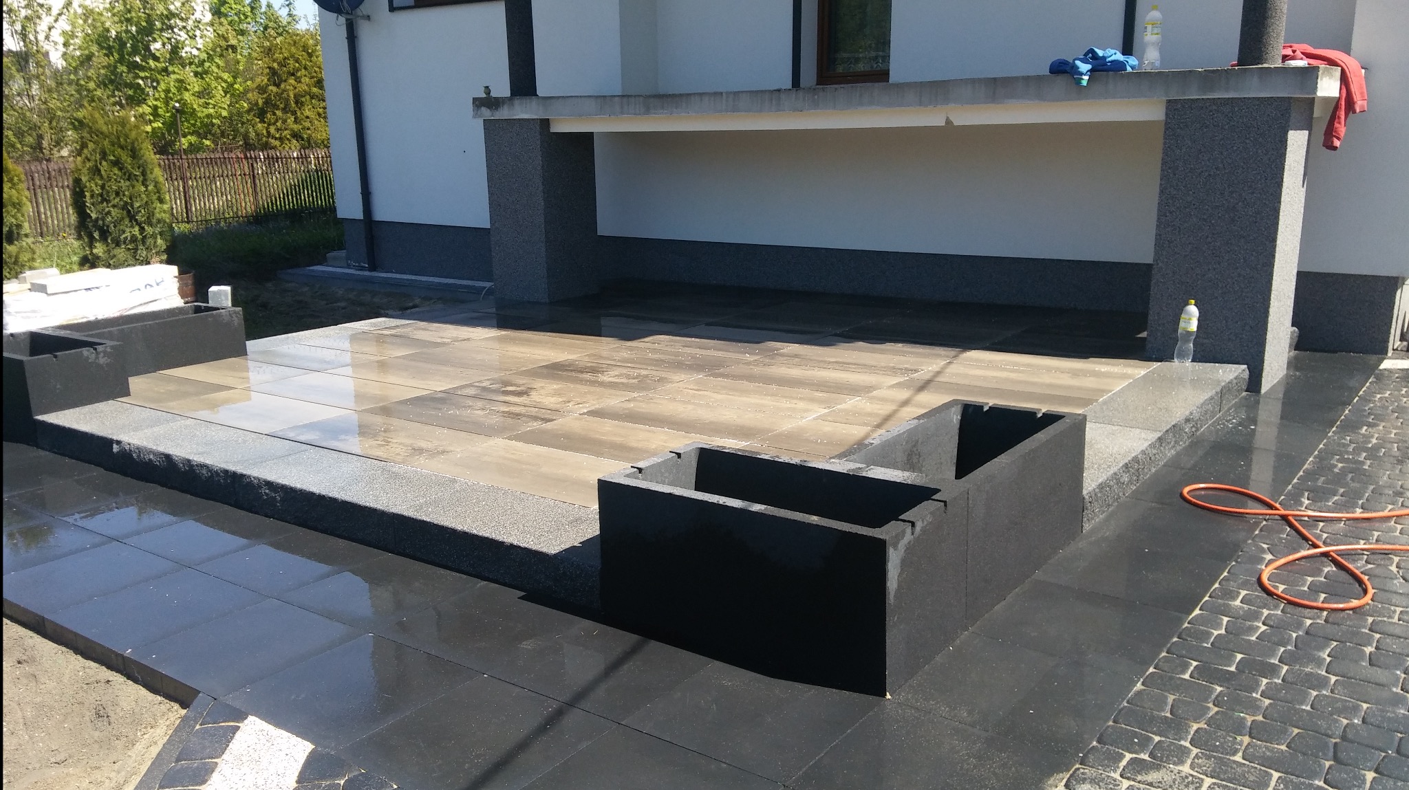 Świeżo ułożona, mokra kostka brukowa w dwóch odcieniach szarości, z nowoczesnymi donicami z betonu architektonicznego i widocznym w tle fragmentem elewacji budynku.