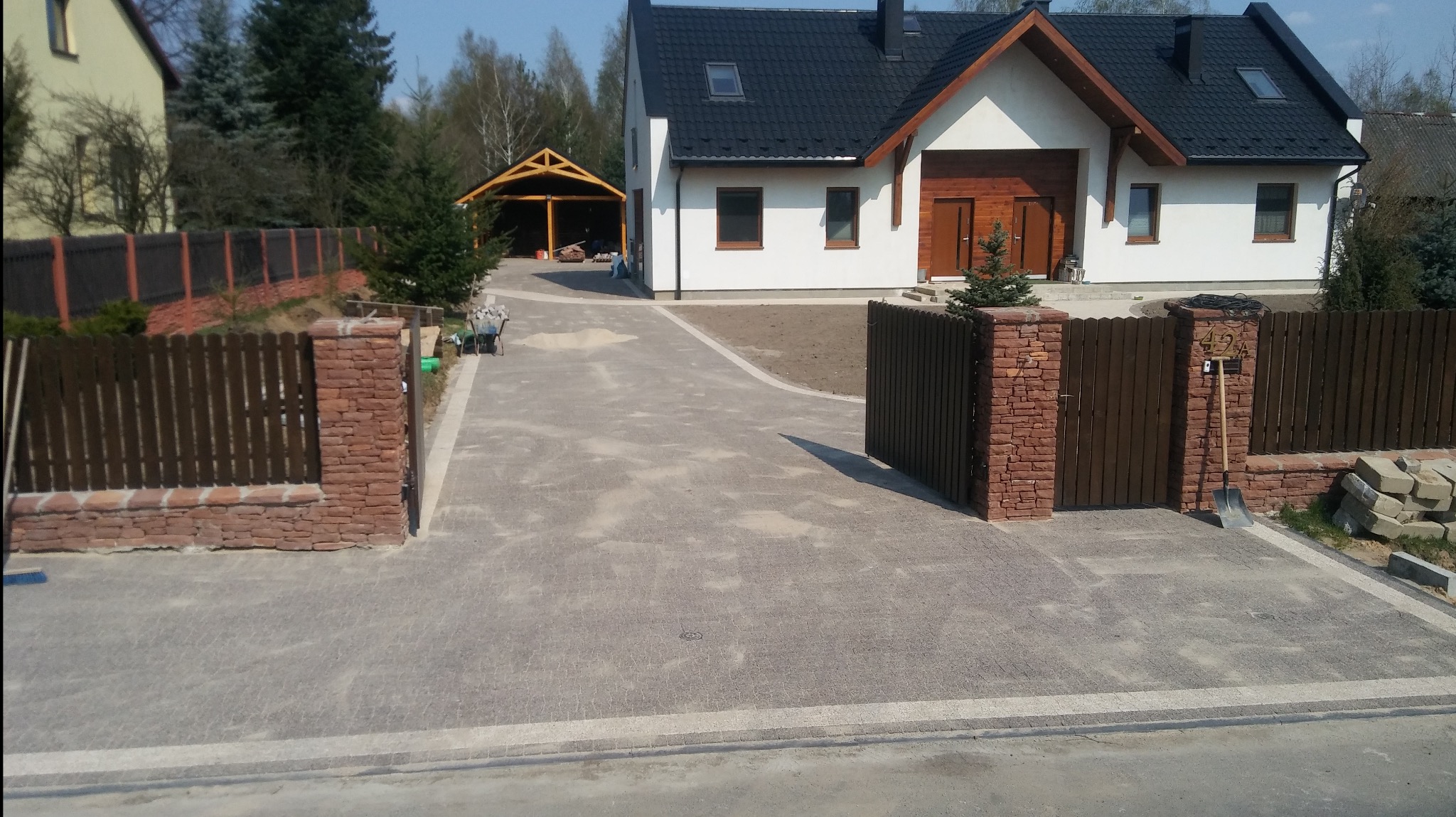 Nowo ułożona, szara kostka brukowa prowadząca do nowoczesnego domu z czarnym dachem i drewnianą bramą wjazdową, widoczne narzędzia i materiały budowlane.