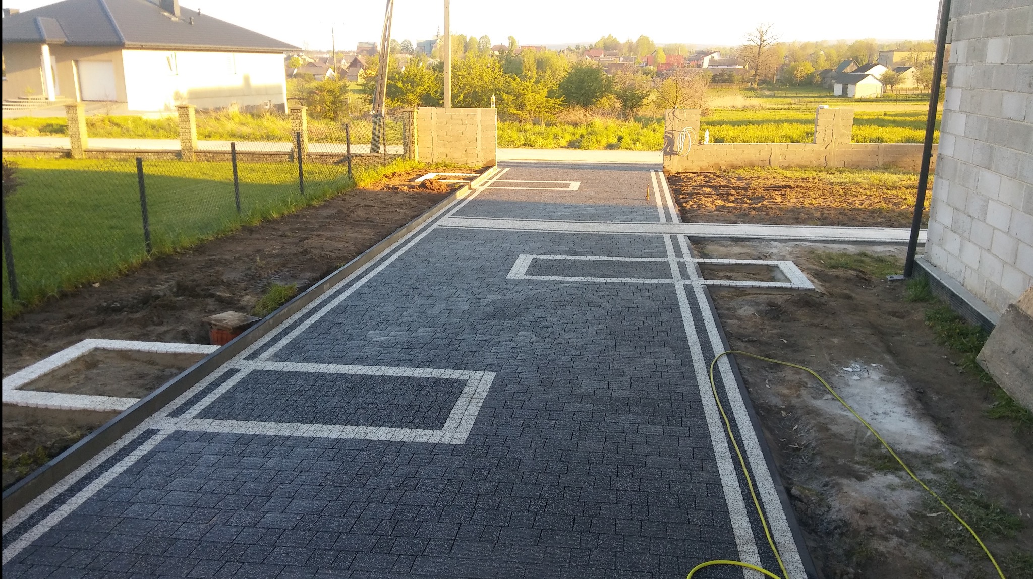 Nowo ułożona, szara kostka brukowa z białymi wstawkami w kształcie prostokątów, obrzeża z kostki, widoczne przygotowane miejsca na zieleń, w tle dom i ogród.