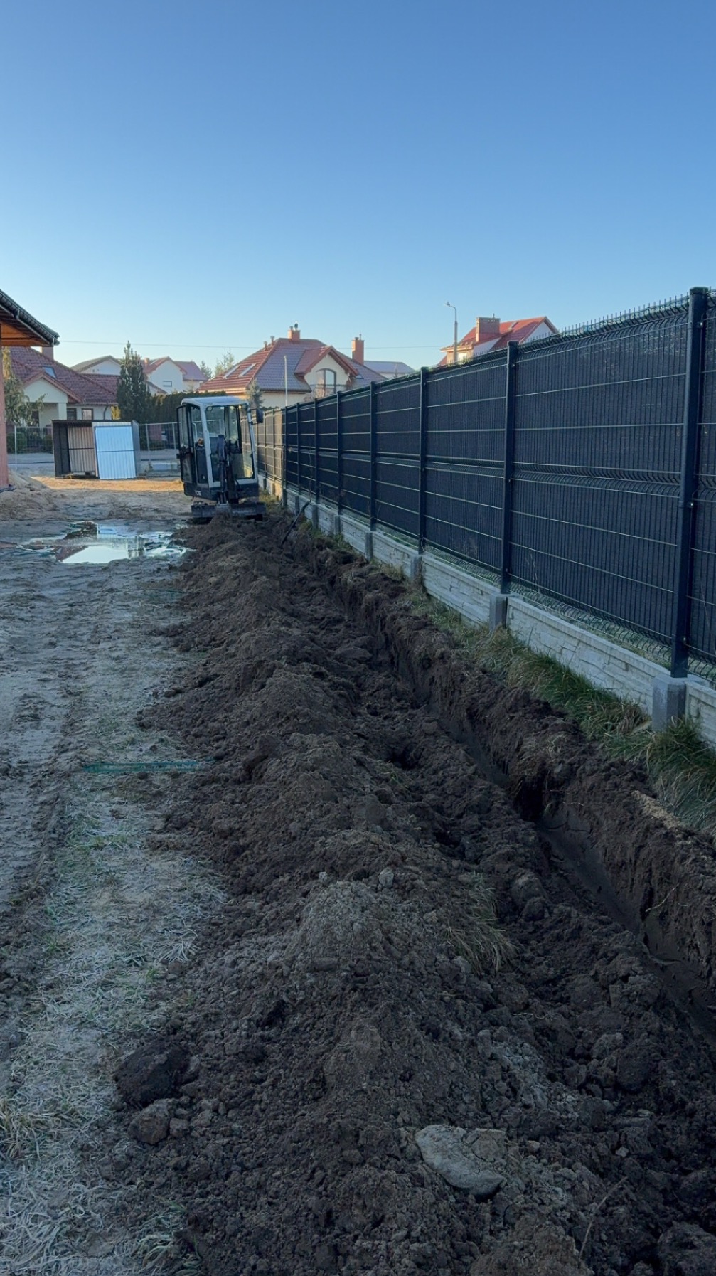 Mini koparka obok wykopu wzdłuż ogrodzenia panelowego na działce budowlanej, widoczne ślady opon i świeżo wykopana ziemia.