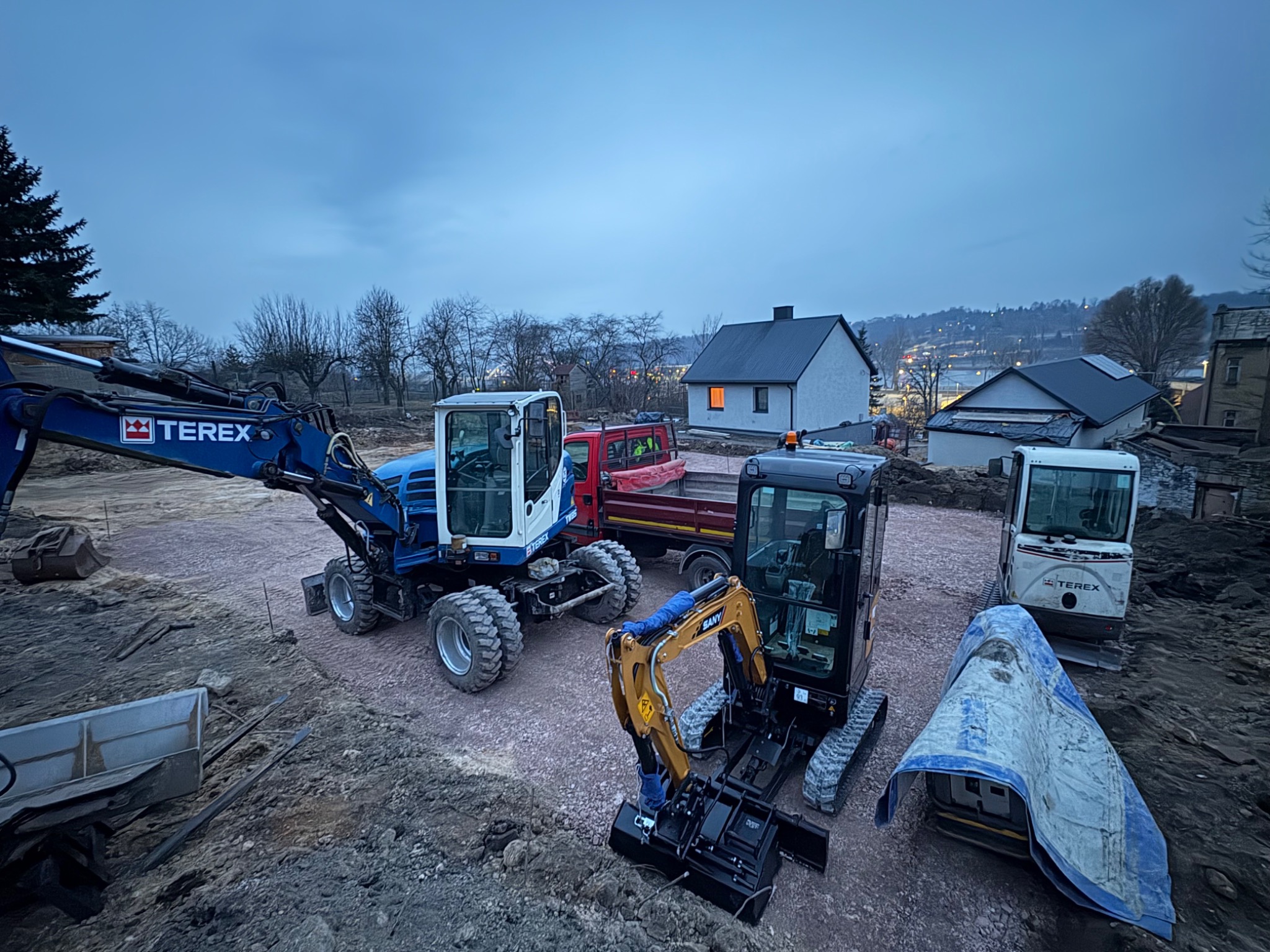 Plac budowy z trzema maszynami: niebieski koparko-ładowarka Terex, mini koparka SANY i czerwona wywrotka w tle, przygotowane do prac ziemnych o zmierzchu.