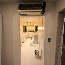 SUNWALL SPÓŁKA Z OGRANICZONĄ ODPOWIEDZIALNOŚCIĄ - Wnętrze w trakcie prac wykończeniowych: widok przez przejście z surowymi ścianami, w tle jasna kuchnia z białymi meblami i wiszącymi lampami, na podłodze wiadro z masą szpachlową i klimatyzator...