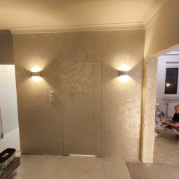 SUNWALL SPÓŁKA Z OGRANICZONĄ ODPOWIEDZIALNOŚCIĄ - Beżowa ściana z dekoracyjną strukturą, oświetlona dwoma kinkietami, z ukrytymi drzwiami i widocznym fragmentem pracującego mężczyzny w tle.