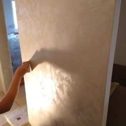 SUNWALL SPÓŁKA Z OGRANICZONĄ ODPOWIEDZIALNOŚCIĄ - Nakładanie dekoracyjnej, beżowo-złotej powłoki na ścianę za pomocą gąbki, widoczne załamania światła, częściowo zakryte meble w tle.