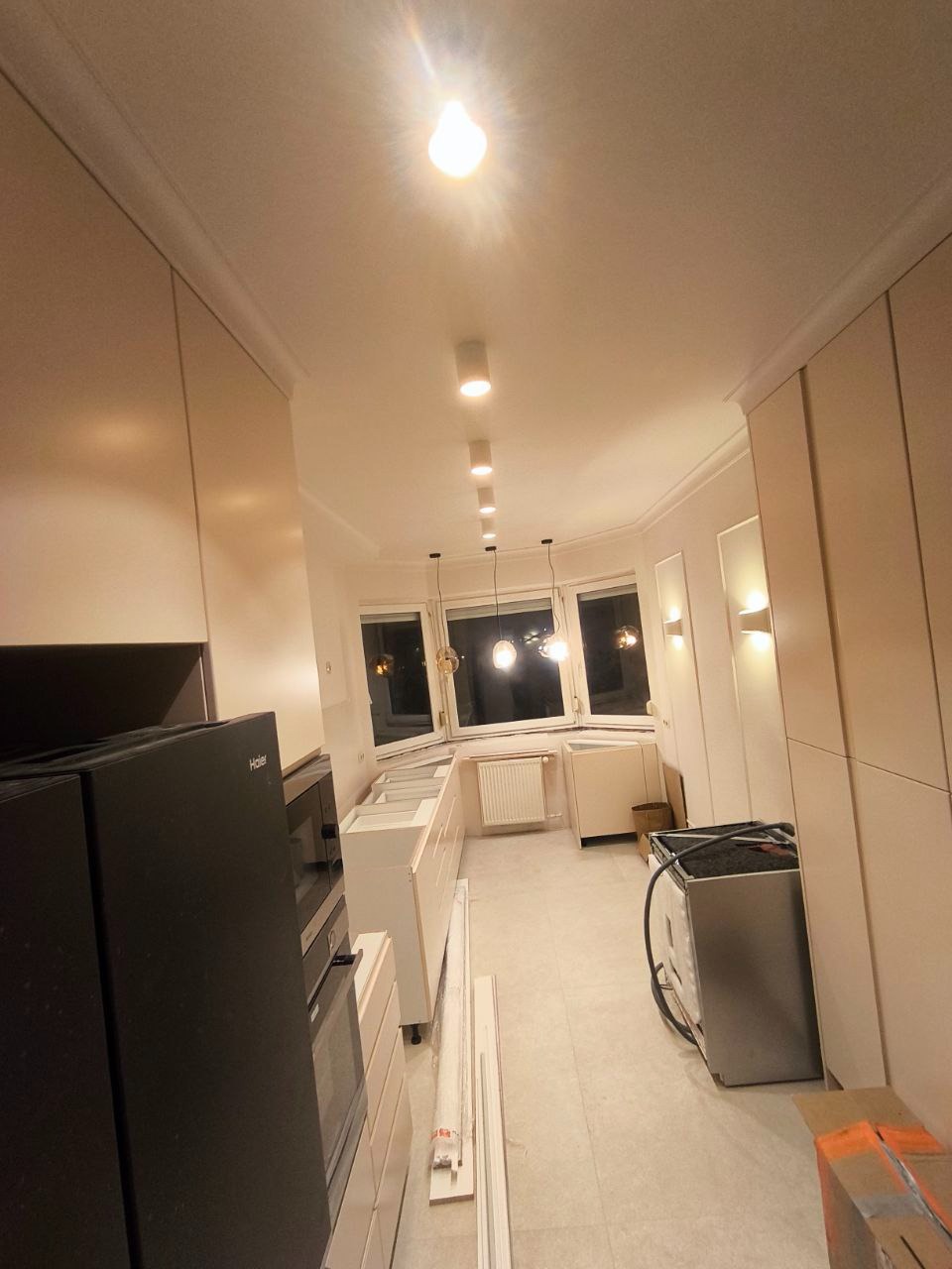 Wnętrze kuchni w trakcie montażu mebli, widoczne niedokończone szafki, sprzęt AGD i elementy wykończeniowe, oświetlenie punktowe sufitowe.